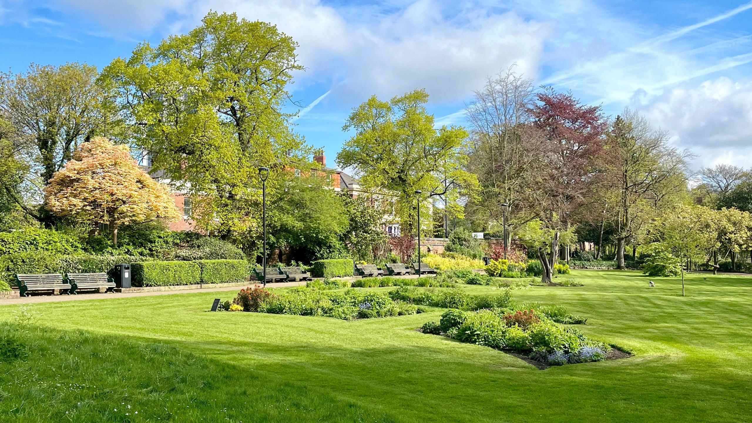 Well-kept lawns and flower beds in Walsall Arboretum, surrounded by mature trees, benches and pathways on a bright spring day.