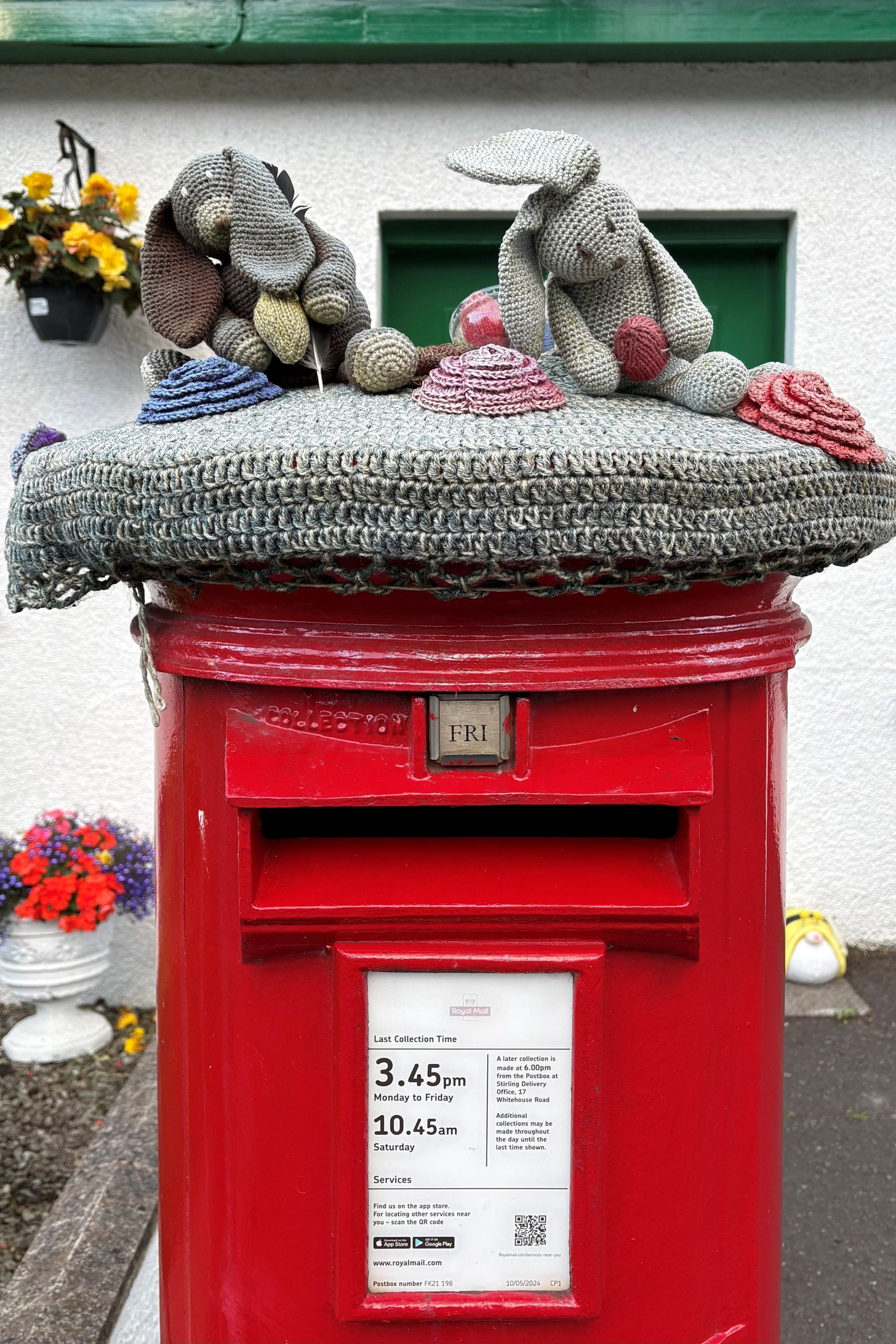 A red post box is topped with a knitted cover featuring a dog and rabbit among colourful yarn flowers, their ears and bodies angled as if being blown sideways by the wind.