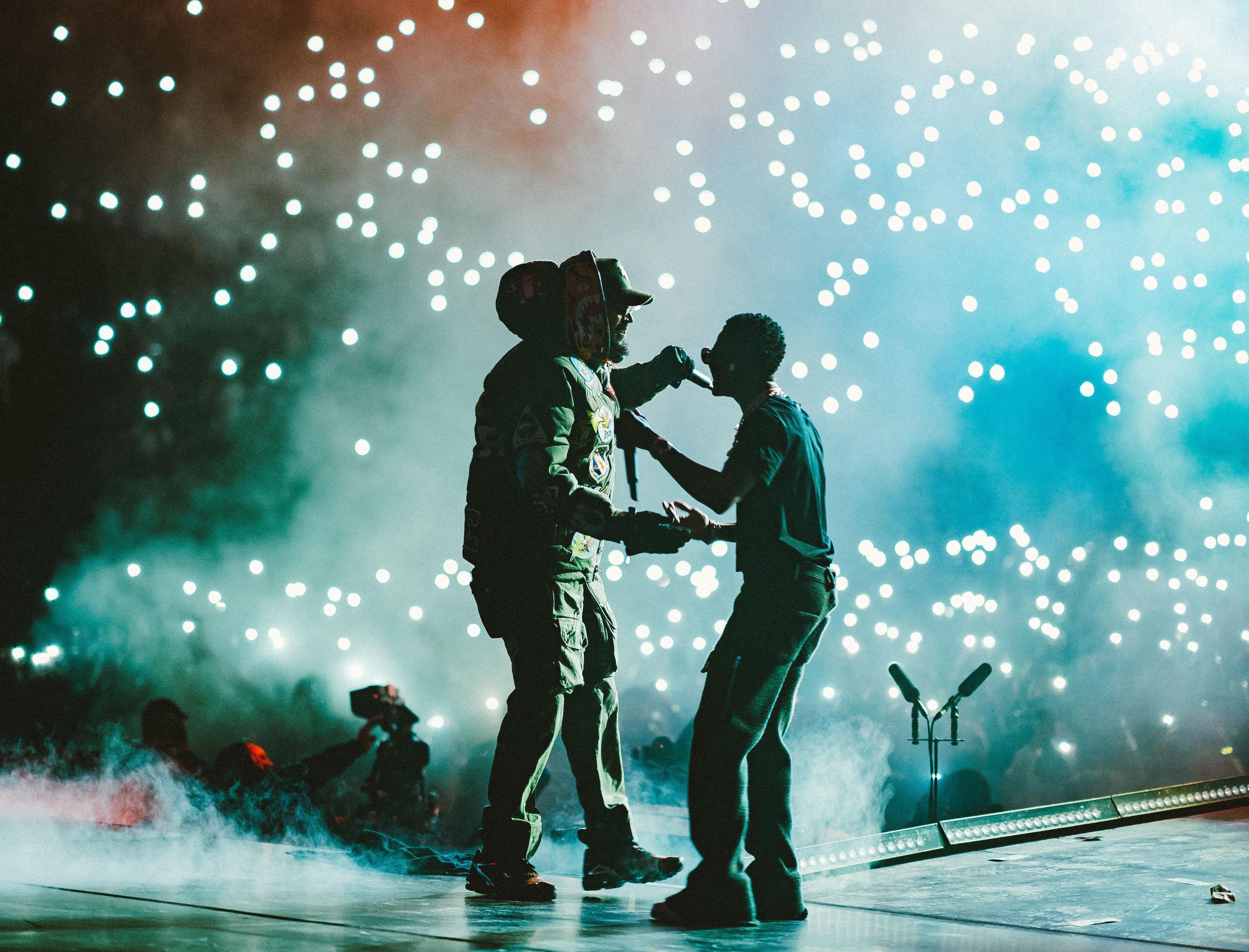 Chris Brown (left) in dark puffer jacked and Wizkid (right) in sunglasses, dark T-shirt and trousers, greet each other on stage at The O2 arena in November 2021. The pair are almost in silhouette as the lights of audience phones sparkle like fairy lights in front of them. Dry ice also lingers in the air.