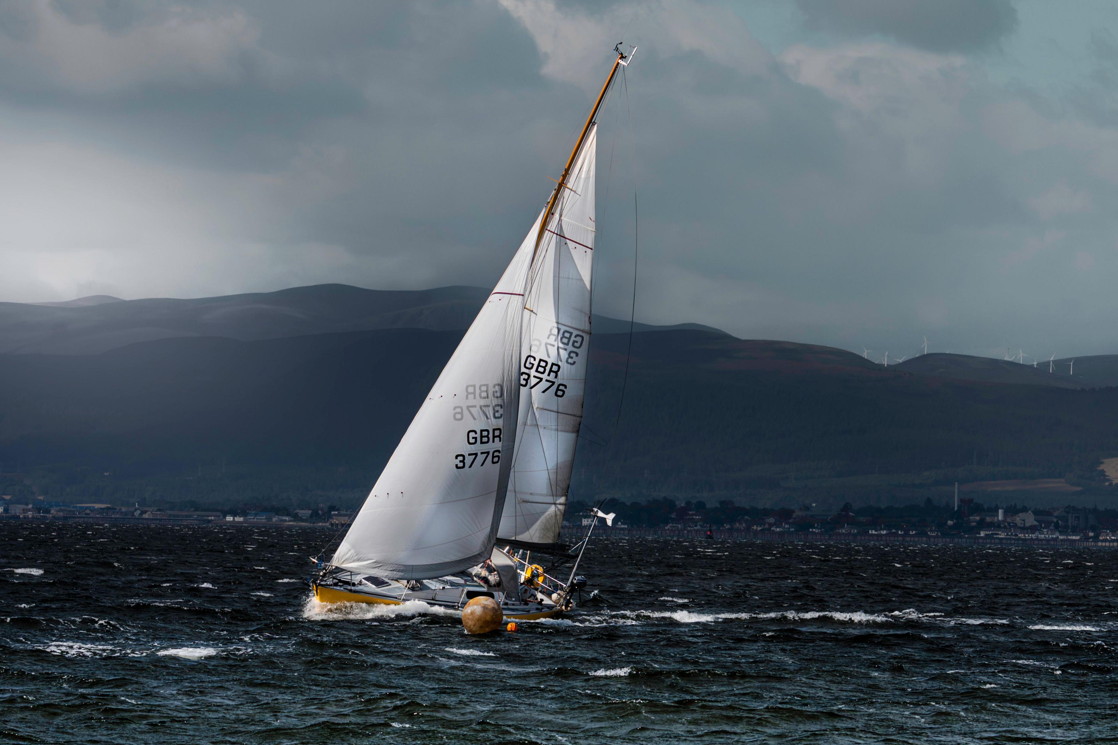 A yacht heels sharply in a strong breeze as it rounds a buoy on choppy water, with dark clouds and the hills above Invergordon in the background.
