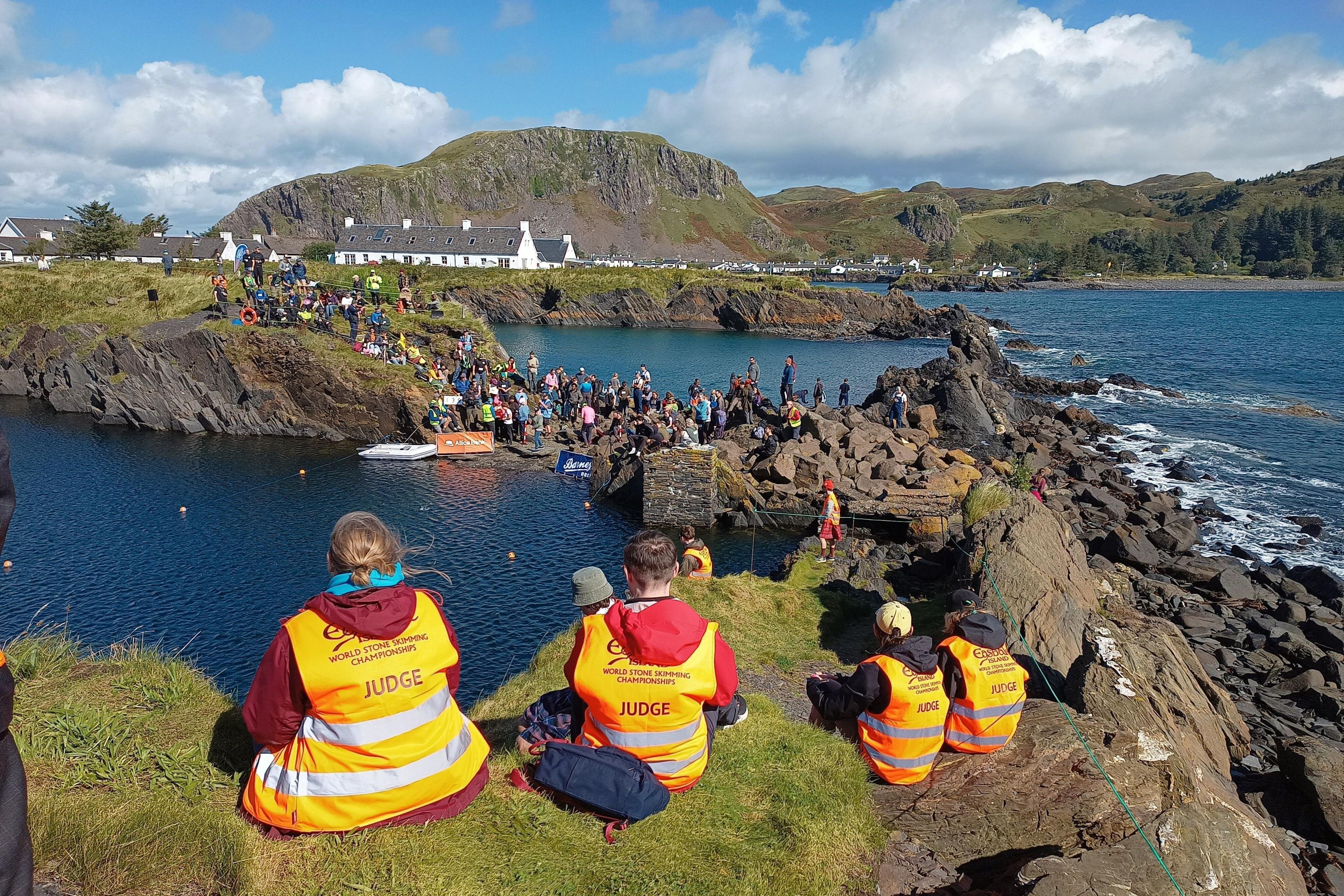 Judges in bright orange vests watch competitors and spectators gathered around a rocky sea pool during the World Stone Skimming Championships, with cottages and the coastline beyond.