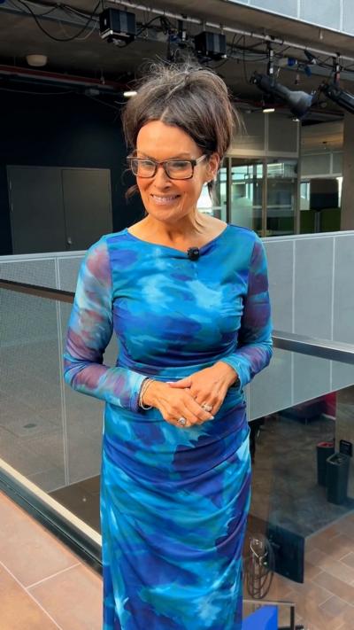 A woman stands smiling at the camera. She has glasses, an updo and blue maxi dress on.