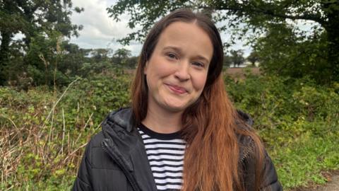 Emma Lewandowicz pictured outside with shrubs and trees in the background. She is wearing a black puffer jacket and black and white striped top and her long brown hair is loose.