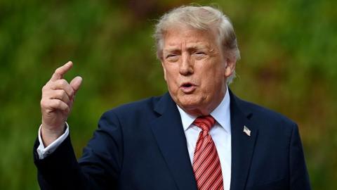 Donald Trump gestures to someone off camera. He is wearing a navy blue suit jacket, white shirt and a reddish-striped tie. 