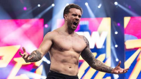 Wrestler Will Ospreay is topless and has his move open as he roars towards a crowd. It's a close cropped photo of him from the waste up. He has both arms outstretched and has a full sleeve tattoo covering all of his left arm.