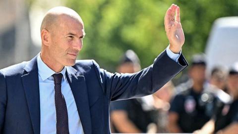 Zinedine Zidane, wearing a dark blue suit, light blue shirt and dark blue tie, waves to crowds after being unveiled as France manager