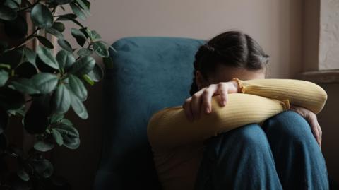 Girl with dark hair has her head hidden in her arms wrapped round her knees sitting on the floor in front of a blue sofa and house plant.