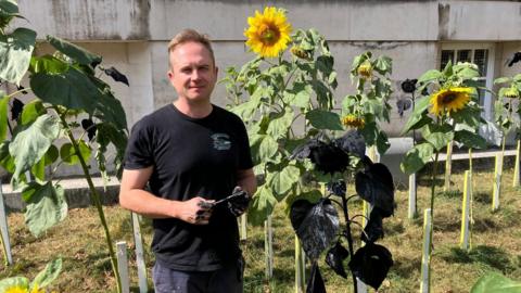 Duncan McKellar stands next to a sunflowers painted black and holding a paintbrush with black paint all over his hands.