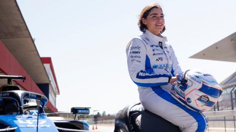 Jamie Chadwick in a white and blue race suit perched on the wheel of a blue F1 car.