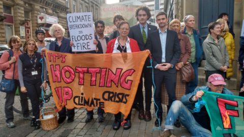 A group of people stood together outside. They are holding an orange banner with pink and black writing on which says: 'It's hotting up climate crisis'. Another banner reads 'climate breakdown floods and fire'.