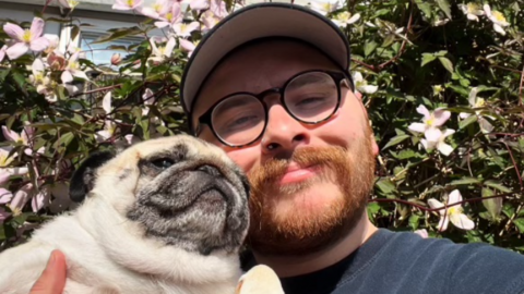 Jack is posing with a small dog in a garden. He is holding the dog close to his face and smiling. He has ginger hair, a beard and a cap on and glasses. There is tree blossom behind him in the sunshine.