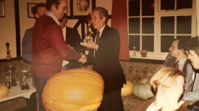 Two men shaking hands and standing behind a giant pumpkin in a pub in a picture taken in the 80s which is slightly grainy and taken on film