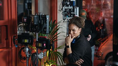 Director Angela Griffin sits on set amongst production equipment smiling at the camera. Her hair is tied up and she wears all black.
