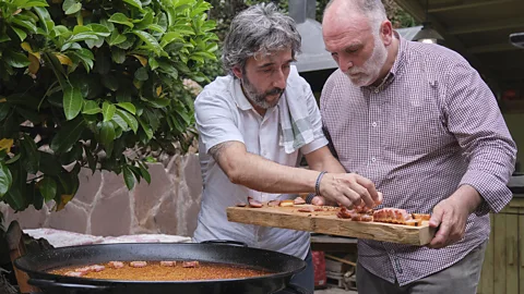 Two men hold a charcuterie board while standing next to a large paella pan (Credit: Courtesy of José Andrés and Family in Spain)