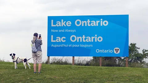 A woman with a dog stood photographing a large blue sign reading "Lake Ontario Now and Always" in English and French (Credit: Getty Images)