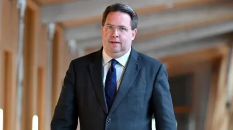 Getty Images A man with dark hair and glasses in a dark blue suit with a blue tie and white shirt walking through the corridors of the Scottish Parliament