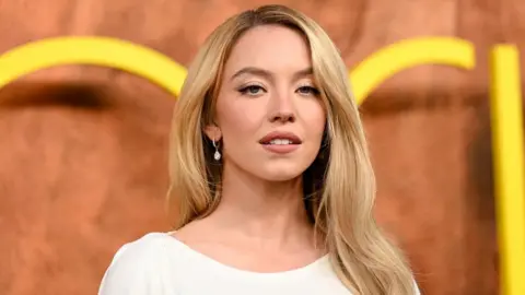 A young woman with long blonde hair wearing a white dress smiles at the camera