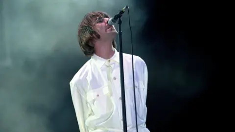 PA Media Liam Gallagher, dressed in white, is lurching into a microphone on a stand as he performs at Knebworth.