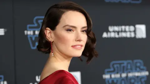 Daisy Ridley poses for photographers with a black wall behind her with Star Wars logs scattered across it. She has brown curly hair and is wearing a red dress.